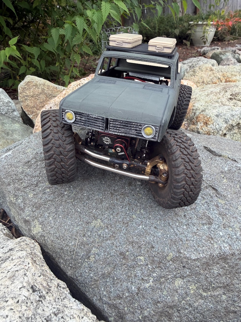 May include: A gray remote-controlled crawler truck featuring large treaded tires, a black roof rack with two beige storage cases, yellow circular headlights, and a visible red winch component mounted on the front bumper assembly.