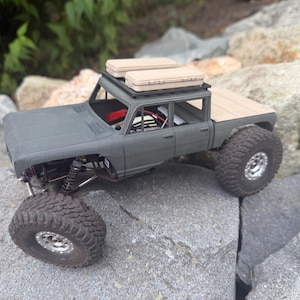 May include: A matte gray remote-controlled crawler truck with a four-door cab, a flatbed, and large knobby tires. The vehicle features a black roof rack carrying two tan rectangular storage cases and an open cabin interior with visible wiring.