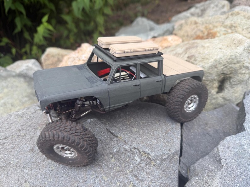 May include: A matte gray remote-controlled crawler truck with a four-door cab, a flatbed, and large knobby tires. The vehicle features a black roof rack carrying two tan rectangular storage cases and an open cabin interior with visible wiring.