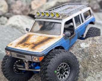 May include: A blue and white Toyota scale model off-road vehicle with a weathered, rusted hood, a roof rack, and five yellow KC auxiliary lights. The truck features large, knobby black tires, a silver front bumper, and visible suspension components.