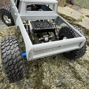 May include: A silver-colored remote-controlled truck model with a visible chassis, black off-road tires, and suspension components. The tailgate features the embossed text INTERNATIONAL. The truck has a black fuel cell and a black M-logo bracket.