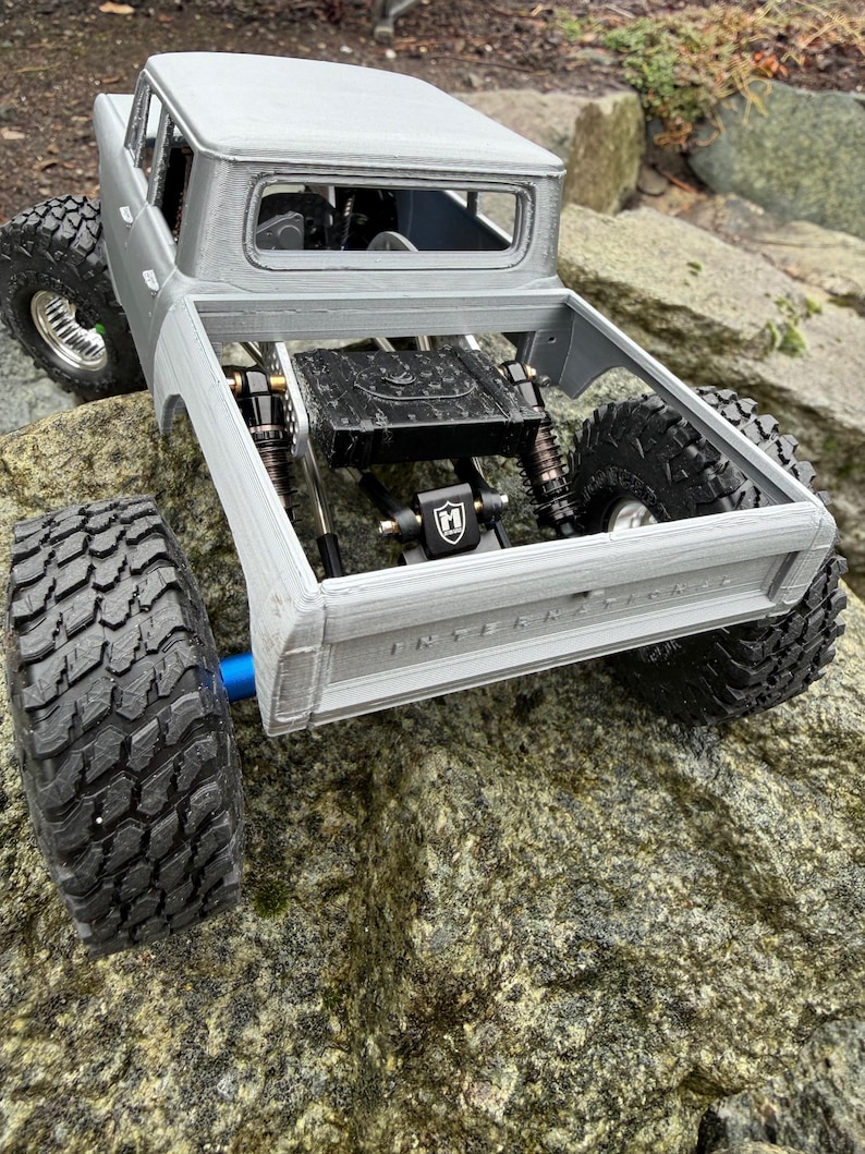 May include: A silver-colored remote-controlled truck model with a visible chassis, black off-road tires, and suspension components. The tailgate features the embossed text INTERNATIONAL. The truck has a black fuel cell and a black M-logo bracket.