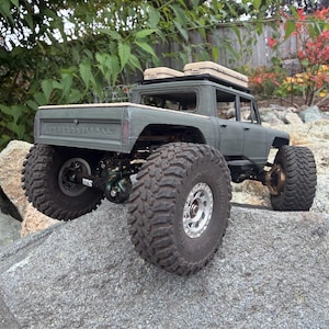 May include: A matte olive green remote-controlled pickup truck with large, deep-tread black off-road tires and silver rims. The truck features a roof rack with two tan storage cases and a detailed rear chassis with visible suspension components.