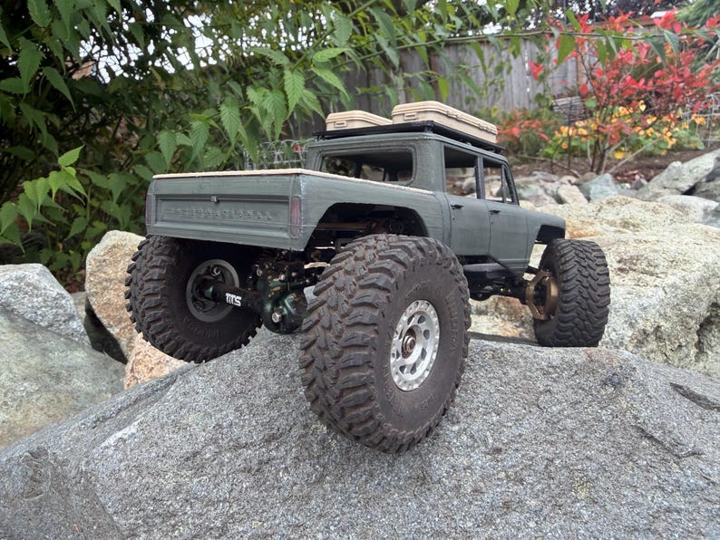 May include: A matte olive green remote-controlled pickup truck with large, deep-tread black off-road tires and silver rims. The truck features a roof rack with two tan storage cases and a detailed rear chassis with visible suspension components.