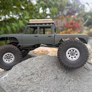 May include: A matte gray scale model pickup truck with a crew cab, large black off-road tires, silver wheels, and a tan storage case mounted on the roof rack. The vehicle features a high-clearance suspension system and a flatbed cargo area.