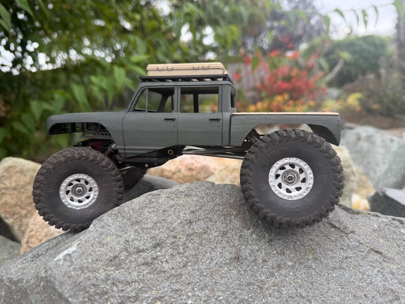 May include: A matte gray scale model pickup truck with a crew cab, large black off-road tires, silver wheels, and a tan storage case mounted on the roof rack. The vehicle features a high-clearance suspension system and a flatbed cargo area.