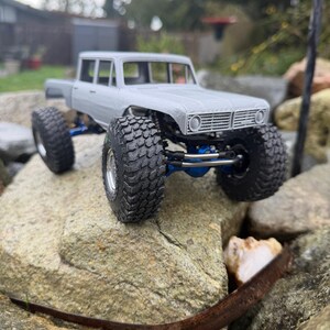 May include: A gray scale model truck featuring a four-door cab, large black off-road tires with deep treads, and a visible blue metal suspension system. The vehicle has a rectangular front grille and a detailed chassis design.
