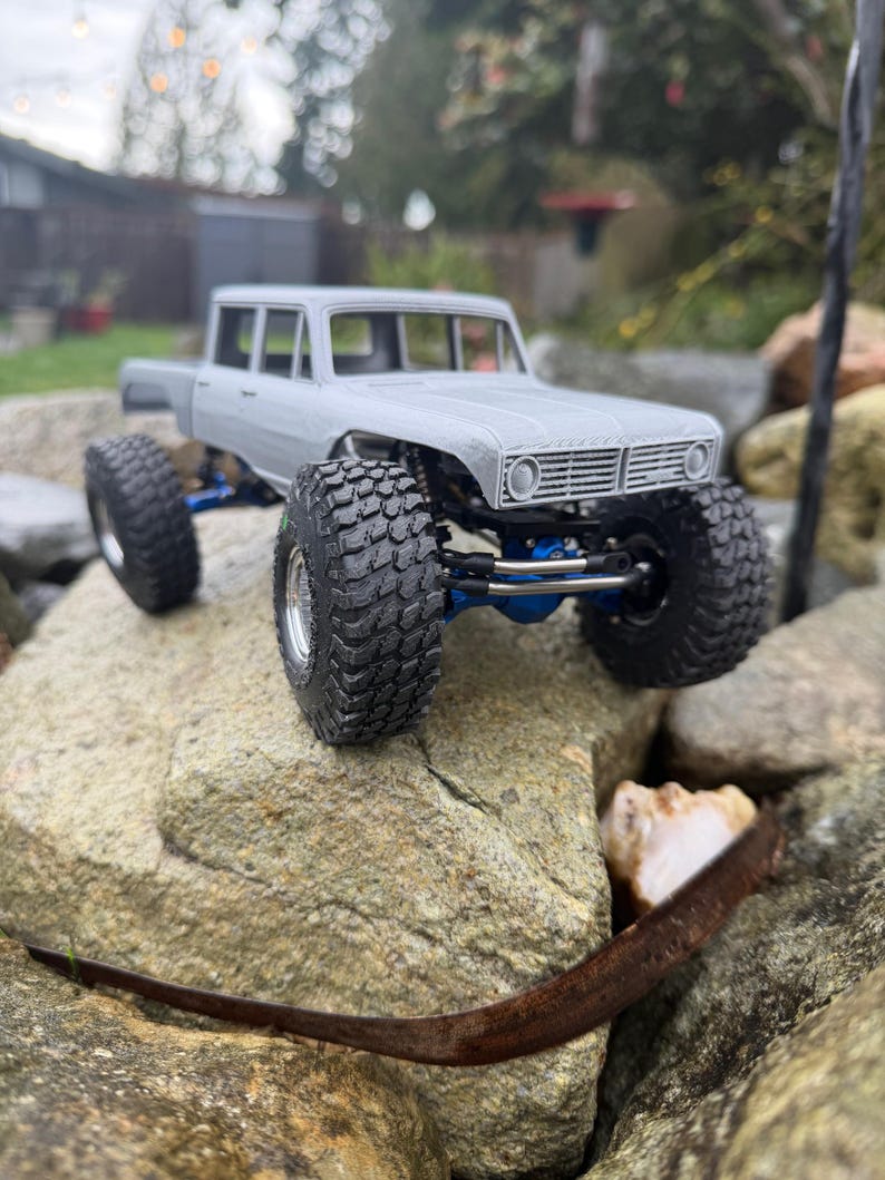 May include: A gray scale model truck featuring a four-door cab, large black off-road tires with deep treads, and a visible blue metal suspension system. The vehicle has a rectangular front grille and a detailed chassis design.