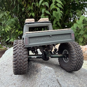 May include: An olive green remote-controlled pickup truck model with large, knobby black tires and a visible suspension system. The tailgate features the embossed text INTERNATIONAL, and the roof rack holds two small tan storage cases.