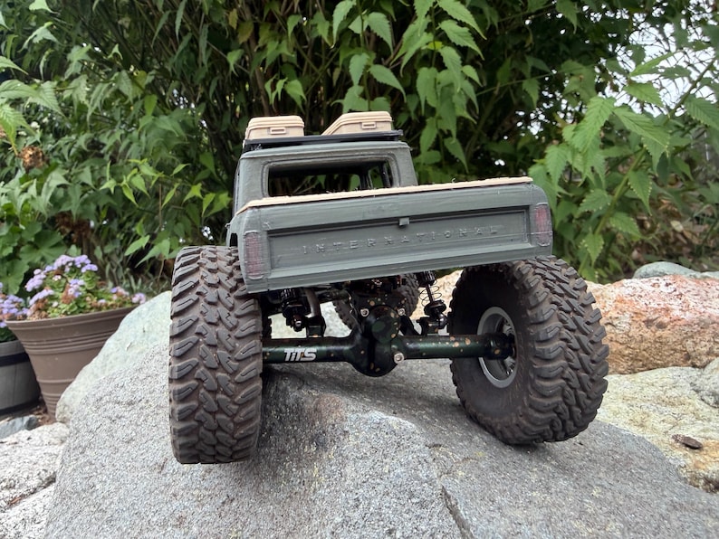 May include: An olive green remote-controlled pickup truck model with large, knobby black tires and a visible suspension system. The tailgate features the embossed text INTERNATIONAL, and the roof rack holds two small tan storage cases.