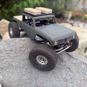 May include: A matte olive green remote-controlled pickup truck model with large black off-road tires, silver rims, and two beige rectangular storage cases secured to a black roof rack. The vehicle features yellow circular headlights.