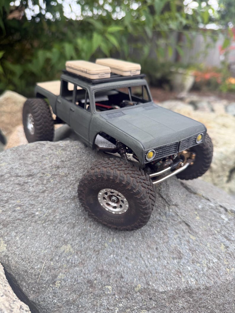 May include: A matte olive green remote-controlled pickup truck model with large black off-road tires, silver rims, and two beige rectangular storage cases secured to a black roof rack. The vehicle features yellow circular headlights.