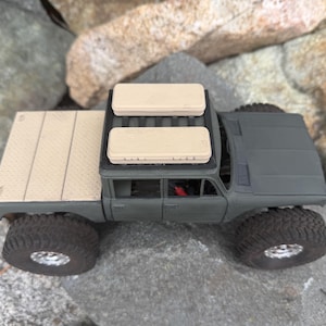 May include: A matte olive green scale model pickup truck featuring a textured tan truck bed cover and two rectangular tan storage cases mounted on the roof rack. The vehicle has large black off-road tires with silver rims.