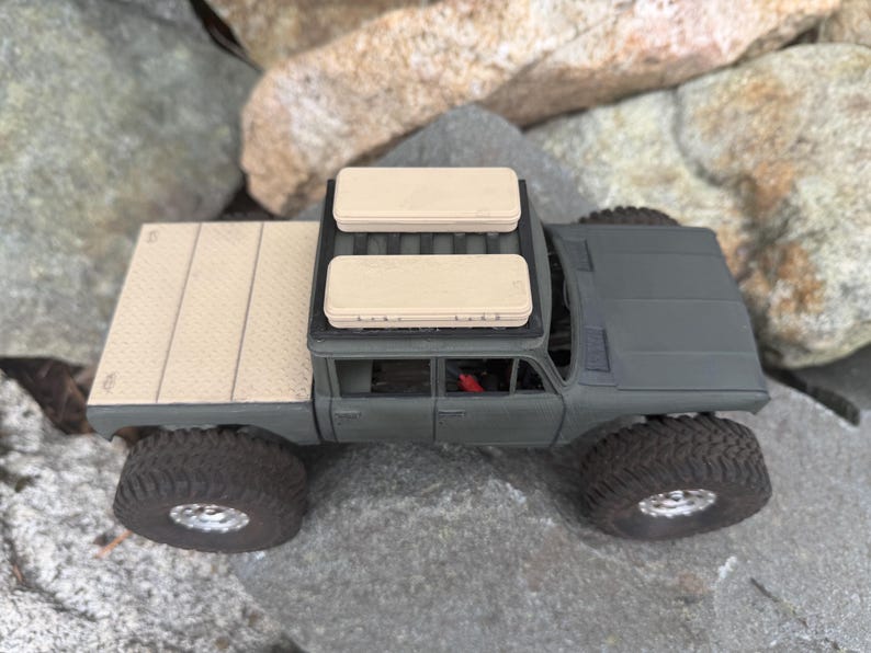 May include: A matte olive green scale model pickup truck featuring a textured tan truck bed cover and two rectangular tan storage cases mounted on the roof rack. The vehicle has large black off-road tires with silver rims.