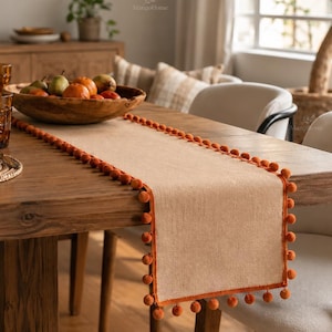 Op de afbeelding: Een rechthoekige beige tafelloper van geweven stof, voorzien van een decoratieve rand met kleine, ronde, roestoranje pompons langs de zijkanten. De loper ligt plat op een houten tafel en dient als ondergrond voor een houten schaal met fruit.