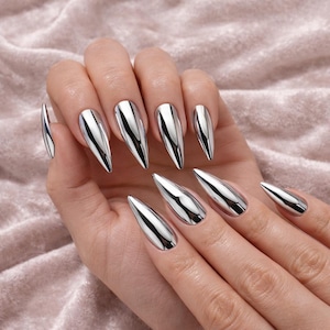 May include: A hand displays a set of silver, metallic, stiletto-shaped artificial nails. The nails have a reflective, mirror-like finish, and are long and pointed. The background is a soft, pinkish-beige fabric.