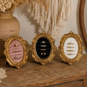 Könnte beinhalten: Drei verzierte, goldgerahmte Ringdisplays. Eines hat einen rosa Samtinnenraum, ein anderes einen schwarzen Samtinnenraum und das dritte einen weißen Innenraum. Jedes enthält mehrere Ringe mit verschiedenen Edelsteinfassungen.