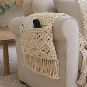 May include: A cream-coloured macrame armrest organiser draped over a sofa arm. The organiser features a woven diamond pattern, a deep pocket containing a white book and a black remote control, and a long fringe trim along the bottom edge.