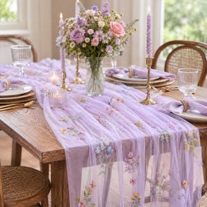 May include: A sheer lavender table runner featuring delicate floral embroidery in shades of pink, yellow, and blue. The runner is draped over a wooden table set with plates, clear ribbed glassware, gold candle holders, and a central flower vase.