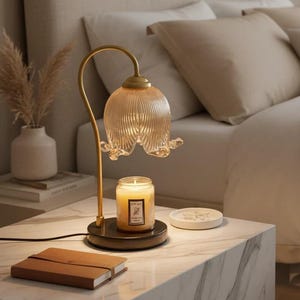 Puede incluir: Una escena de dormitorio con una lámpara dorada con una pantalla de vidrio en forma de flor, una vela encendida y una mesita de noche con tapa de mármol. La cama tiene ropa de cama y almohadas beige. Un cuaderno marrón está sobre la mesa. La lámpara mide unos 30 cm de alto.