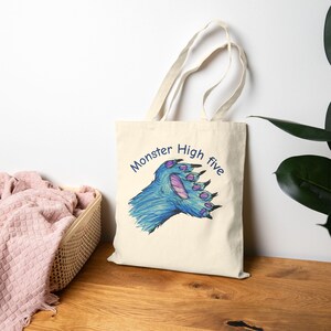 May include: Cream-colored tote bag featuring a blue monster paw with purple accents, giving a high five. The words "Monster High five" are written in blue script above the paw. The bag has a simple design with long handles.
