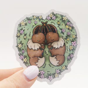 Peut inclure: Un autocollant représentant deux personnages de dessins animés avec une fourrure brune, des cols blancs et des queues touffues, blottis l'un contre l'autre. Ils sont entourés d'un arrangement en forme de cœur d'herbe verte et de fleurs colorées. L'autocollant est tenu par une main.