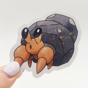 May include: A holographic sticker of a cartoon hermit crab. The crab has orange claws and body, with large black eyes. Its shell is dark grey with a sparkling, iridescent finish. The sticker is held by a person's hand.