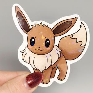 May include: A holographic sticker of Eevee, a brown and cream-coloured Pokémon with large ears and a fluffy tail. The sticker has a glittery, star-like effect. The sticker is held against a grey background.