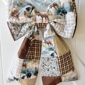 May include: A large, decorative bow made of quilted fabric. The bow features a patchwork design with various patterns, including moose, bears, and landscape scenes. The color palette includes shades of brown, beige, and white. A light blue ribbon is attached for hanging.