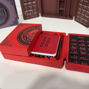 Pu&ograve; includere: Una scatola organizer per giochi rettangolare rossa con un design circolare nero sul coperchio. La scatola aperta contiene un taccuino Field Notes rosso, una penna nera e un pannello di tracciamento delle abilit&agrave; con testo nero su sfondo rosso.