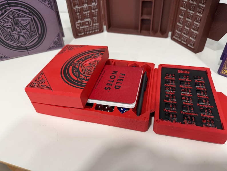 Pu&ograve; includere: Una scatola organizer per giochi rettangolare rossa con un design circolare nero sul coperchio. La scatola aperta contiene un taccuino Field Notes rosso, una penna nera e un pannello di tracciamento delle abilit&agrave; con testo nero su sfondo rosso.