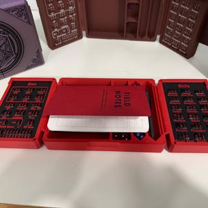 Pu&ograve; includere: Un organizer da tavolo rosso a tre ante con uno scomparto centrale contenente un taccuino Field Notes e una penna nera. I pannelli laterali presentano inserti neri con testo rosso per statistiche e abilit&agrave;, incluse le etichette Stats, Skills e Death Saves.
