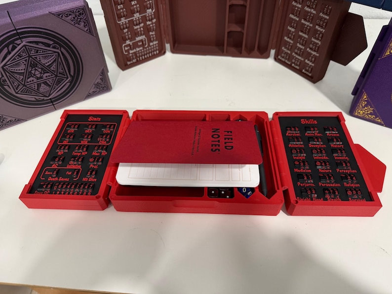 Pu&ograve; includere: Un organizer da tavolo rosso a tre ante con uno scomparto centrale contenente un taccuino Field Notes e una penna nera. I pannelli laterali presentano inserti neri con testo rosso per statistiche e abilit&agrave;, incluse le etichette Stats, Skills e Death Saves.