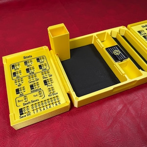 Pu&ograve; includere: Un organizer da tavolo in plastica giallo brillante con tracker meccanici integrati per le statistiche e le abilit&agrave; del personaggio. L'unit&agrave; include uno scomparto centrale foderato in nero, un portadadi verticale e quadranti numerici rotanti etichettati con testo come Stats, HP, AC e Skills.