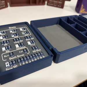 Pu&ograve; includere: Una custodia rettangolare blu scuro per il tracciamento di giochi con coperchio incernierato. L'interno presenta uno scomparto foderato in feltro grigio e un pannello di tracciamento meccanico con quadranti numerici rotanti per statistiche e HP.