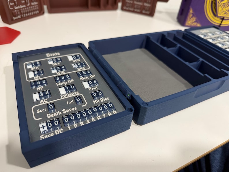 Pu&ograve; includere: Una custodia rettangolare blu scuro per il tracciamento di giochi con coperchio incernierato. L'interno presenta uno scomparto foderato in feltro grigio e un pannello di tracciamento meccanico con quadranti numerici rotanti per statistiche e HP.