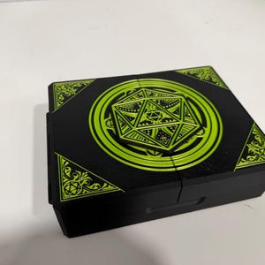 Pu&ograve; includere: Una scatola rettangolare nera con una grafica verde lime brillante sul coperchio. Il design include un'icona centrale di un dado a venti facce circondata da motivi circolari e decorazioni angolari con piccoli disegni di dadi.