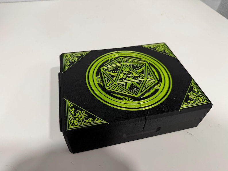 Pu&ograve; includere: Una scatola rettangolare nera con una grafica verde lime brillante sul coperchio. Il design include un'icona centrale di un dado a venti facce circondata da motivi circolari e decorazioni angolari con piccoli disegni di dadi.