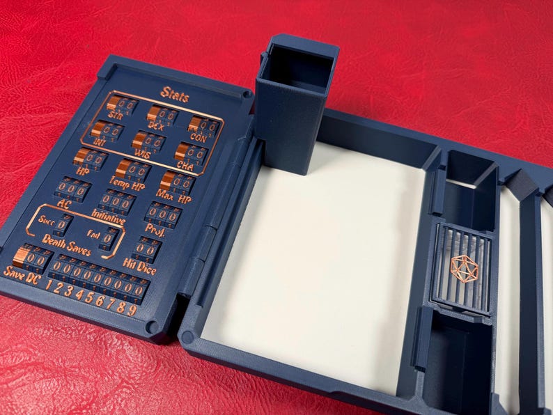 Pu&ograve; includere: Un organizer da tavolo in plastica blu scuro con un tracker meccanico delle statistiche integrato, numeri ed etichette color rame che includono Stats, STR, DEX, CON, INT, WIS, CHA, HP, Temp HP, Max HP, AC, Initiative, Proj, Death Saves e Hit Dice.
