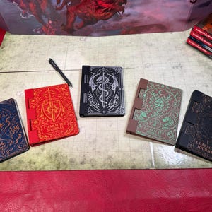 May include: Five character journals with unique embossed designs, including a sword, dragon, and ornate patterns. The journals are in various colors, such as red, blue, and brown, with the words "Character Journal" on the cover. A black pen rests nearby.