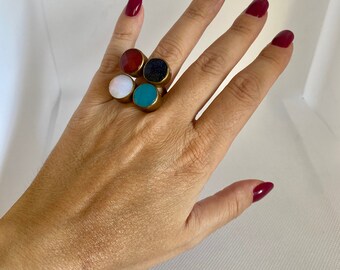 Könnte beinhalten: Ein goldener Ring mit fünf verschiedenen farbigen Steinen. Die Steine sind rot, weiß, blau, schwarz und türkis.