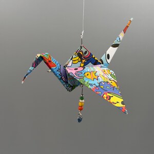 May include: A hanging origami crane folded from paper with a colourful underwater scene featuring yellow fish, a pink octopus, and a scuba diver. A small silver wire loop at the top holds a dangling charm with four multi-coloured beads and a blue crystal.