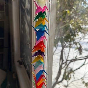 May include: A string of colorful origami cranes, suspended in front of a window. The cranes are made of folded paper in various colors, including green, pink, blue, and orange. The window is in the background, with a view of trees and a bright sky.