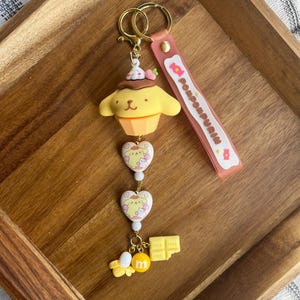 May include: A yellow and brown keychain featuring a cartoon character in a cupcake design, with a pink strap that says "Pompompurin". The keychain has gold-coloured hardware and includes heart-shaped charms and a chocolate bar charm.