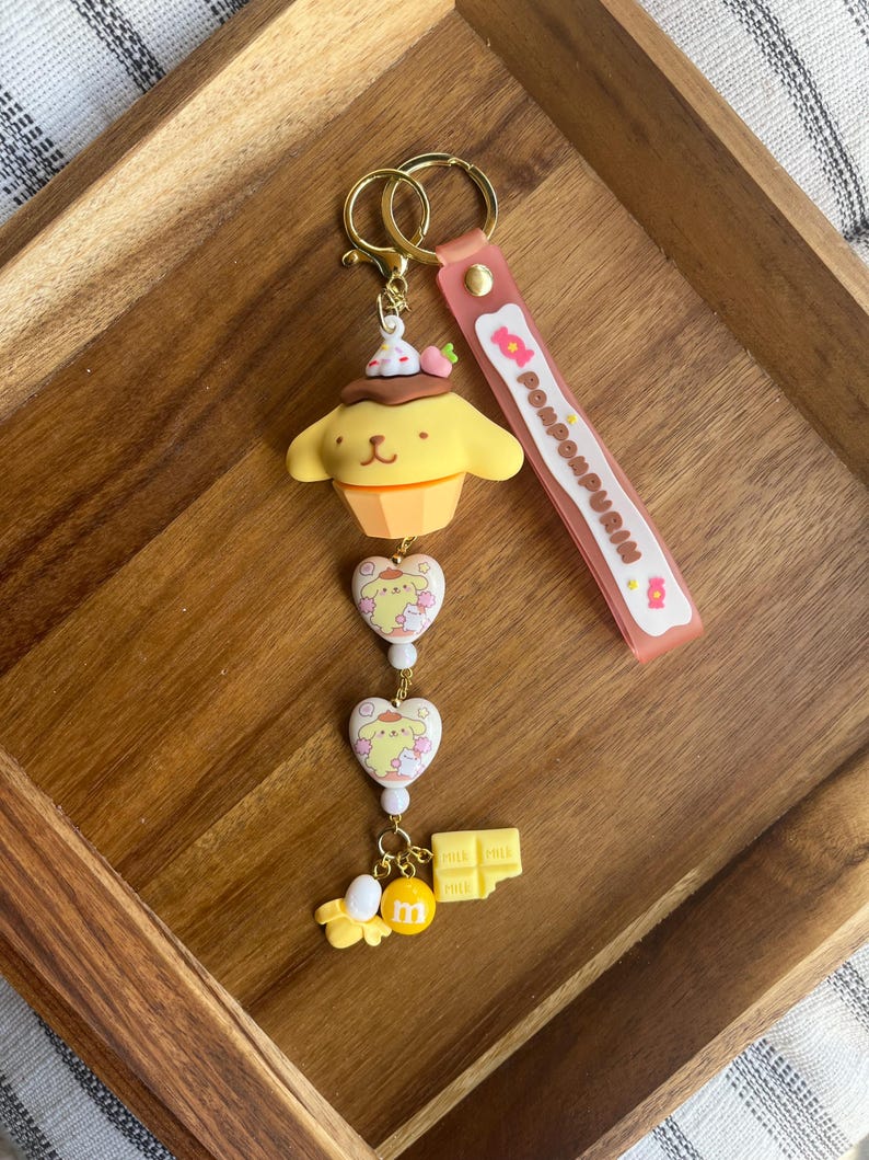 May include: A yellow and brown keychain featuring a cartoon character in a cupcake design, with a pink strap that says "Pompompurin". The keychain has gold-coloured hardware and includes heart-shaped charms and a chocolate bar charm.