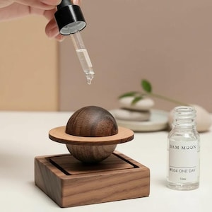 Puede incluir: Un difusor de aceites esenciales de madera con forma de planeta con un cuentagotas dispensando líquido. El difusor se encuentra sobre una base cuadrada, y junto a él hay un pequeño frasco de vidrio con una etiqueta. La etiqueta dice "3AM MOON #004 ONE DAY 10ml."