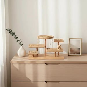 May include: A tiered wooden jewelry organizer with multiple levels and small trays. The organizer is displayed on a light-colored dresser, with a white vase and framed picture in the background. Necklaces and earrings are displayed.