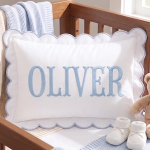May include: A white rectangular pillow with a scalloped edge trimmed in light blue. The name OLIVER is embroidered in large, light blue serif letters across the center of the pillow. The pillow is positioned in a wooden crib with blankets.