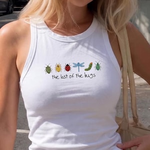 May include: A white sleeveless ribbed tank top featuring a graphic row of six colorful insects including a beetle, ladybug, dragonfly, and caterpillar, with the text "the last of the bugs" printed in a black handwritten font below the insects.