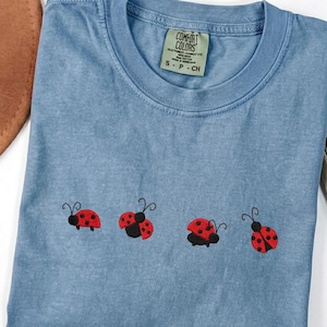 May include: A blue Comfort Colors t-shirt with four embroidered ladybugs in a row. Each ladybug is red with black spots and a black head. The shirt has a small green tag near the collar that says "Comfort Colors".